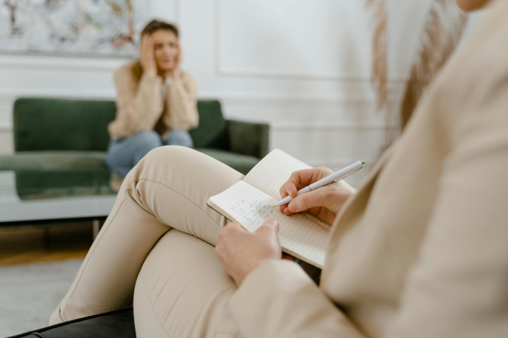 Aus der Perspektive eines Coaches zeigt das Bild im Vordergrund eine Person, die sich Notizen in ein liniertes Notizbuch schreibt. Im unscharfen Hintergrund sitzt eine Frau auf einem grünen Sofa und stützt nachdenklich den Kopf in die Hände.